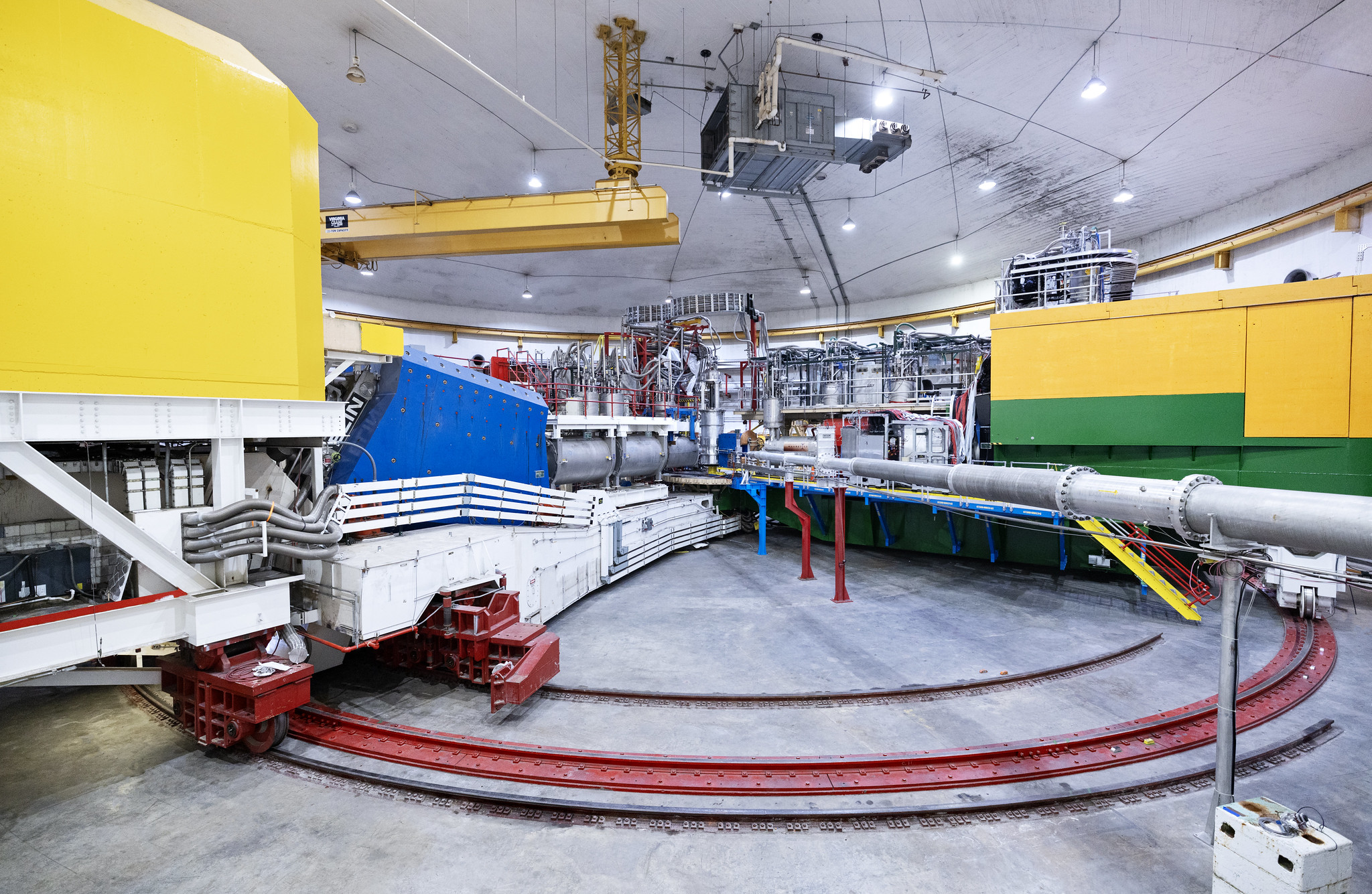 A pair of large particle detectors, one painted yellow and blue and the other painted yellow and green, rest inside the large, drum-shaped Experimental Hall C at Jefferson Lab. A steel beampipe is in teh foreground and the detectors are connected to wires, pipes, conduits and supports. 